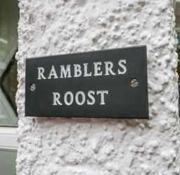 A black sign with white letters saying Ramblers Roost mounted on a textured white wall outside a building at Ramblers Roost in Grasmere