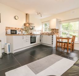 A kitchen with white cabinets and tiled backsplash a wooden dining table with two chairs and a black leather sofa at Ramblers Roost in Grasmere