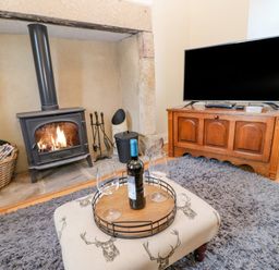 A living room with a wood burning stove basket with logs a wooden cabinet with a television and a tray with a wine bottle and glasses on a cushioned stool at East Farmhouse Cottage in Humshaugh near Hexham