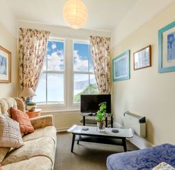 A small living room with a beige sofa and patterned blue chair facing a TV on a stand by a window with floral curtains at Clooneavin Apartment 5 in Lynmouth near Lynton