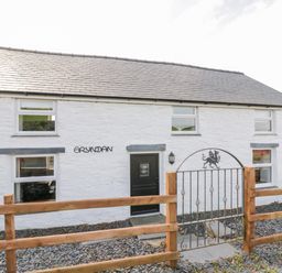 Exterior of a white stone house with black door and windows behind a wooden fence with metal gate at Bryndan in Machynlleth