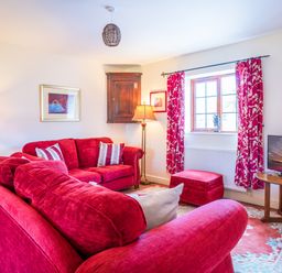 A living room with red sofas a wooden table with a television floral curtains and a window at Nyth Y Wennol in Abersoch