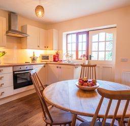 A kitchen with round wooden table and chairs a bowl of apples oven microwave flowers and a door with window at Nyth Y Wennol in Abersoch