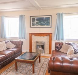 A living room with two brown leather sofas a glass coffee table a fireplace and two windows with blue curtains at 11 Pendarves St Merryn near Padstow