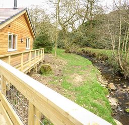 A wooden cabin with a railing deck next to a small stream and trees at Park Brook Retreat in Scorton