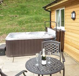 An outdoor patio with a hot tub next to a wooden building and a table with two chairs a bottle and two glasses at Park Brook Retreat in Scorton
