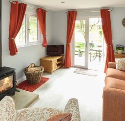 A living room with a wood-burning stove a wicker basket with firewood a television on a wooden stand orange curtains a wooden sideboard a table lamp and an orange sofa at Park Brook Retreat in Scorton
