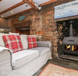 A living room with a beige sofa and red plaid cushions near a wood burning stove in a stone fireplace at Tinners Cottage in Liskeard