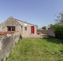 A stone house with a red door a grass lawn and a patio with outdoor furniture at The Stone Barn in Evercreech near Shepton Mallet