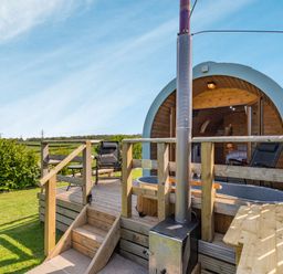 An outdoor wooden deck with chairs a hot tub and a round wooden cabin in a grassy area at Two Hoots in Northop