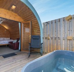 A wooden pod bedroom open to a deck with a hot tub and chair at Two Hoots in Northop