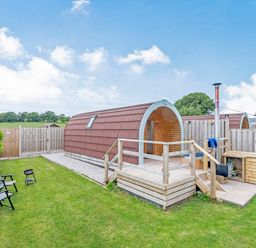 A wooden pod with a deck and chimney in a fenced grassy garden with chairs and a small grill at Ewe Two in Oakenholt near Northop