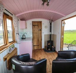 A small room with two armchairs a wood stove a kitchenette and windows with floral curtains at Llety'r Bugail in Holyhead
