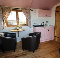 A small kitchen area with pink cabinets and a kettle next to two black armchairs and a wooden table under a window at Llety'r Bugail in Holyhead