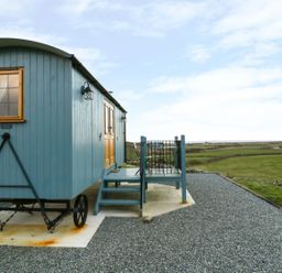 A blue wooden caravan with stairs and railings on a gravel area with green fields in the background at Llety'r Bugail in Holyhead