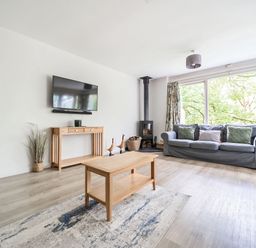 A living room with a gray sofa, wooden coffee table, wall-mounted television, wood-burning stove, and large windows at Cherry - Woodland Cottages in Bowness-in-Windermere