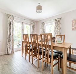 A dining room with a wooden table and eight chairs next to windows with curtains at Cherry - Woodland Cottages in Bowness-in-Windermere