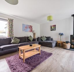 A living room with a gray sectional sofa green cushions wooden coffee table purple rug and a wood stove at Maple - Woodland Cottages in Bowness-on-Windermere