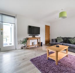 A living room with a gray sofa with cushions a wooden coffee table on a purple rug a mounted flat screen TV on a wooden console and glass doors at Maple - Woodland Cottages in Bowness-on-Windermere
