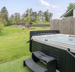 An outdoor hot tub with steps next to a wooden fence and a large grassy area with trees and a wooden cabin in the background at Maple - Woodland Cottages in Bowness-on-Windermere