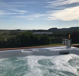 A hot tub with a bottle in an ice bucket and two glasses on a wooden railing overlooking a countryside landscape with hills and wind turbines at Rhos Helyg in Penysarn near Amlwch