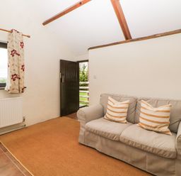A room with a beige sofa and two striped cushions near a window and an open door at Nestling Barn in Peter Tavy near Tavistock