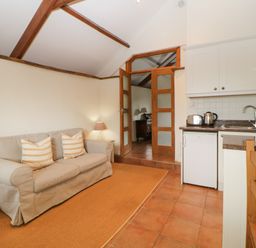 A living room with a beige sofa and striped cushions next to a small kitchen with white cabinets and wooden furniture at Nestling Barn in Peter Tavy near Tavistock