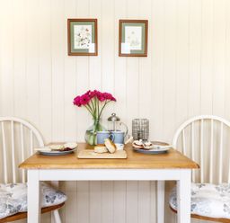 A small wooden table with two white chairs a vase with flowers a cutting board with scones and plates with jam and cream on a wooden wall at Bramble Burlawn near Wadebridge