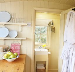 A kitchen counter with a bowl of green apples plates glasses and a sign next to a doorway with a white bathrobe hanging and a small bathroom with a sink and window at Willow Burlawn near Wadebridge