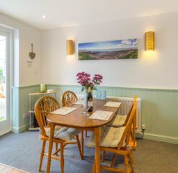 A dining area with a wooden table and four chairs with seat cushions and placemats flowers in a vase and a bottle on the table at Estuary View in Warkworth