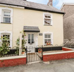 The front of a cream colored cottage with white framed windows a black metal gate and flower pots at Brickfield Cottage in Machynlleth