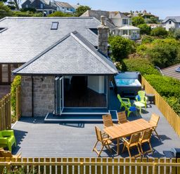 An outdoor deck with wooden dining table and chairs green plastic chairs a hot tub and barbecue at Rocky Close in St Ives