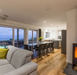 An open living space with a beige sofa with colorful pillows a dining table with chairs a wood stove and large windows overlooking a patio at Chy Lowen in St Ives