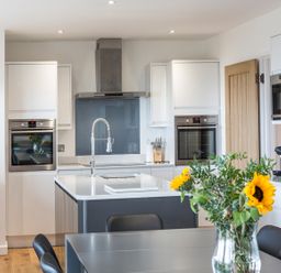 A modern kitchen with white cabinets, two built-in ovens, a kitchen island with a faucet, and a dining table with a vase of sunflowers at Chy Lowen in St Ives