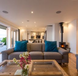 A living room with a gray sofa with blue pillows and a glass coffee table with flowers and an open view to a kitchen and dining area at Boscarne in St Ives