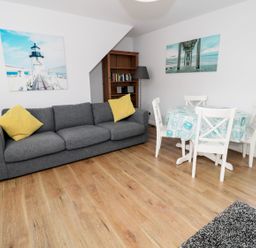 A living area with a grey sofa with yellow cushions a wooden floor a round dining table with four chairs and two wall pictures at Sunnie Cottage in Seahouses