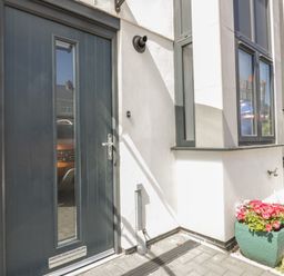 A black front door with a glass panel next to a white wall and windows with a green planter holding pink and white flowers at 5 Ocean Heights in Newquay