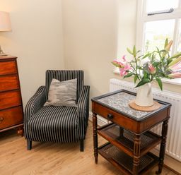 A corner with a striped armchair next to a wooden chest of drawers and a side table with a vase of flowers by a window at Waingate Cottage in Flookburgh