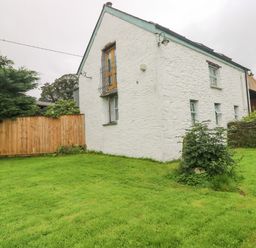 A white stone house with blue trim windows and a green lawn surrounded by wooden fence and trees at Y Cartws in Nevern near Cardigan