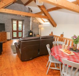 A living room with a brown leather sofa wooden beams a small dining table with a red checkered tablecloth and a wood stove at Y Cartws in Nevern near Cardigan