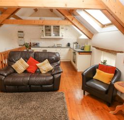 A living area with a brown leather sofa and armchair with cushions a wooden side table with a lamp and a kitchen with white cabinets and wooden beams on the ceiling at Y Cartws in Nevern near Cardigan