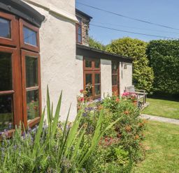 A garden with flowers and green plants outside a white house with wooden framed windows and a bench on a stone pathway at Meadow Lea in Pensilva near Liskeard