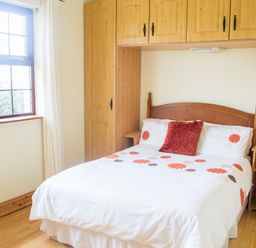 A bedroom with a double bed under wooden built-in cupboards and a window with white curtains at Flynns Cottage in Quilty County Clare