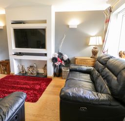 A living room with black leather sofas a red rug a wall mounted television and a wooden cabinet with a lamp at 6 Stad Yr Ysgol in Manod near Blaenau Ffestiniog