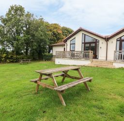 A grassy lawn with two wooden picnic tables in front of a building with large windows and a tiled roof at Keepers Cottage in Sidmouth