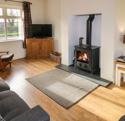 A living room with a wood burning stove a gray sofa a wooden armchair a wooden cabinet with a television and a window with patterned curtains at Rosies Cottage in Bushmills County Antrim