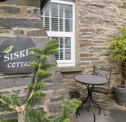 A small outdoor seating area with a round metal table and two chairs next to a stone wall with a window and a sign reading Siskin Cottage at Siskin in Dinas Mawddwy near Dolgellau