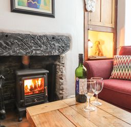 A fireplace with a wood stove burning and fire tools next to a wooden table with a bottle of wine and two glasses beside a red sofa with a patterned cushion at Siskin in Dinas Mawddwy near Dolgellau