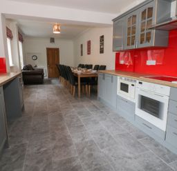 A long kitchen with grey cabinets and red backsplash a wooden dining table with black chairs and a brown leather armchair at Dumbuie in Haltwhistle