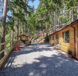 An outdoor area with a wooden cabin on one side a gravel ground and a fenced forest at Lady Bagots Cabin in Ruthin